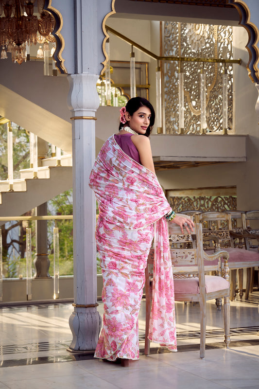 Woman in a white saree with a pink floral print standing near a chair.