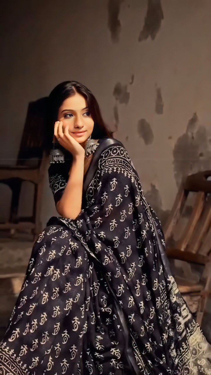 Woman in black and white printed cotton saree with jewelry sitting indoors.