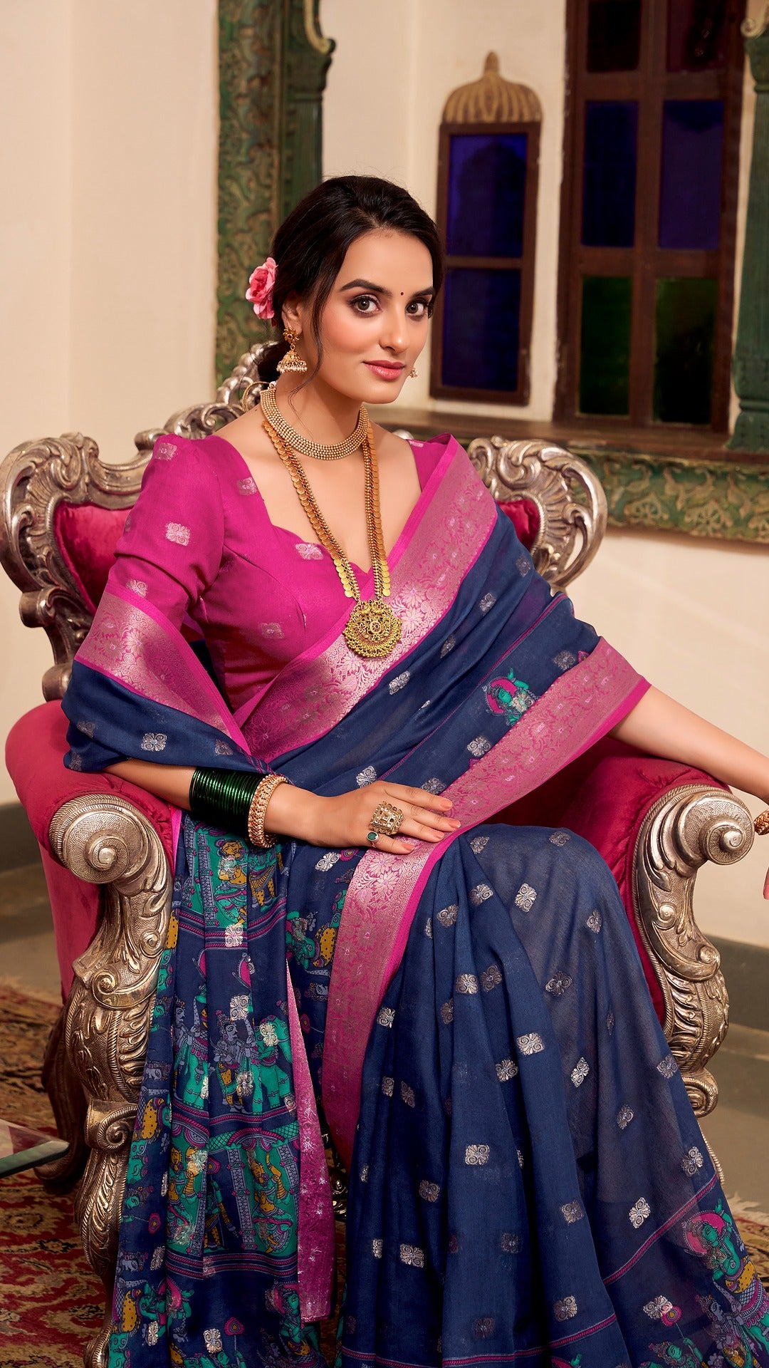 Model sitting in chair wearing navy and pink silk saree with jewelry.