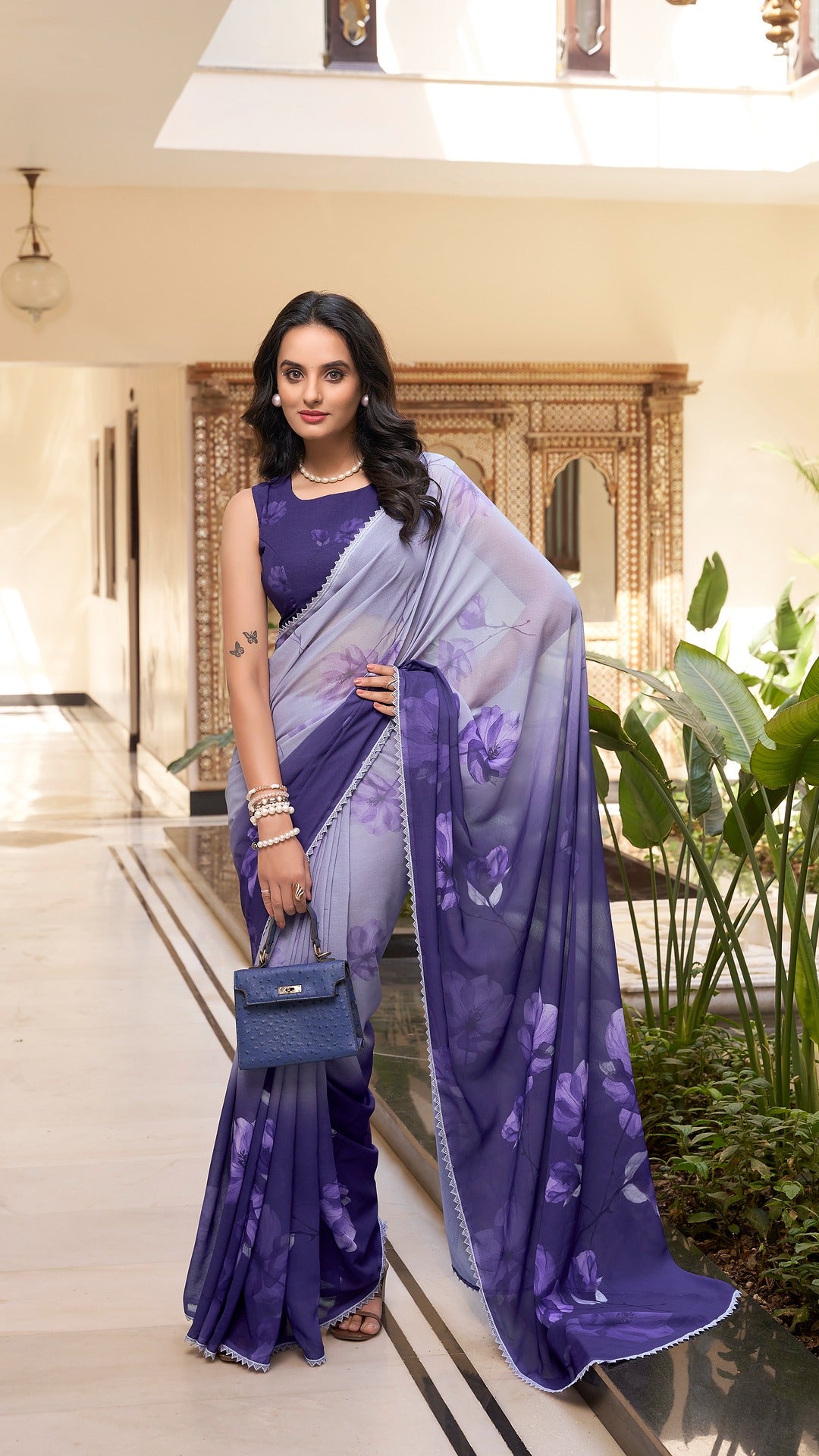 Woman in a stylish purple floral saree holding a blue handbag.