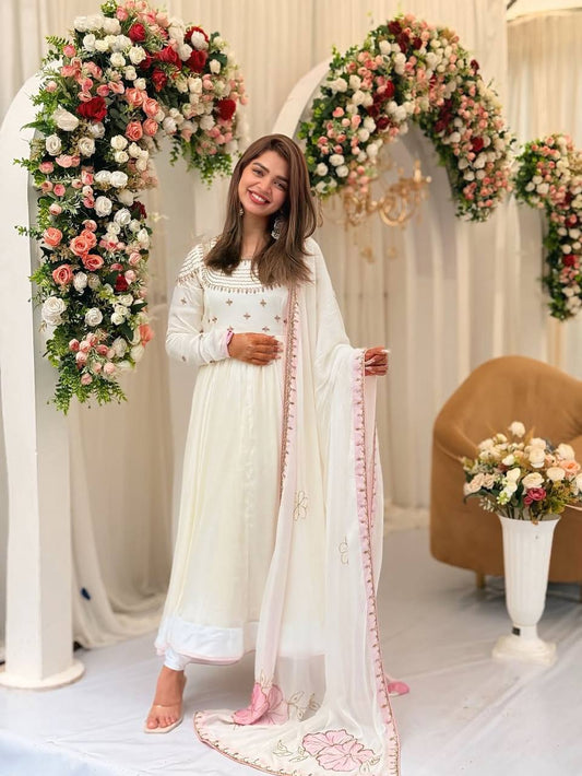 Woman wearing off-white embroidered party gown with pastel pink floral dupatta, standing by floral arch.