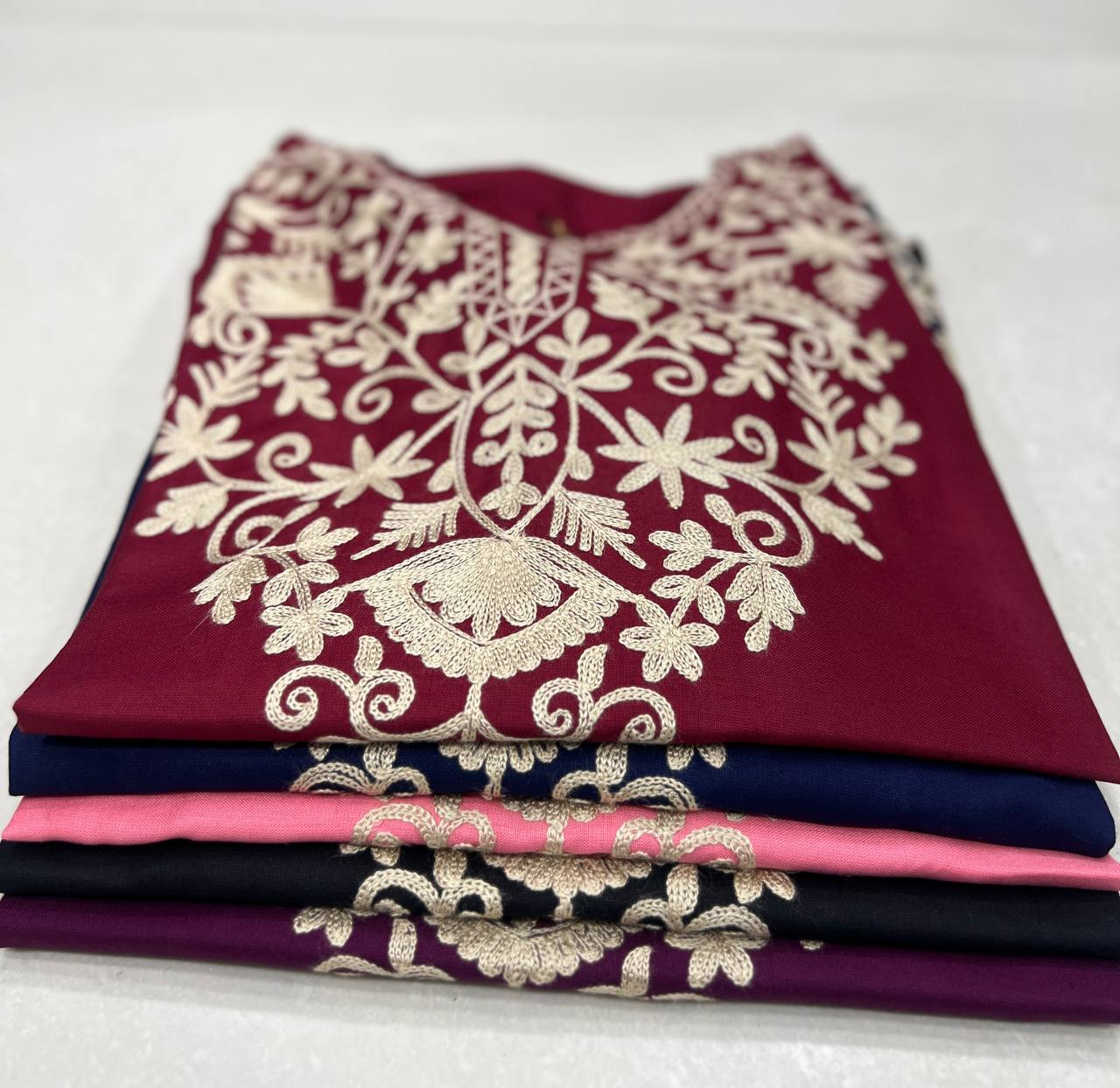 Stack of folded Adaa ShortKurtis in maroon, blue, pink, black, and purple, all with signature floral embroidery.