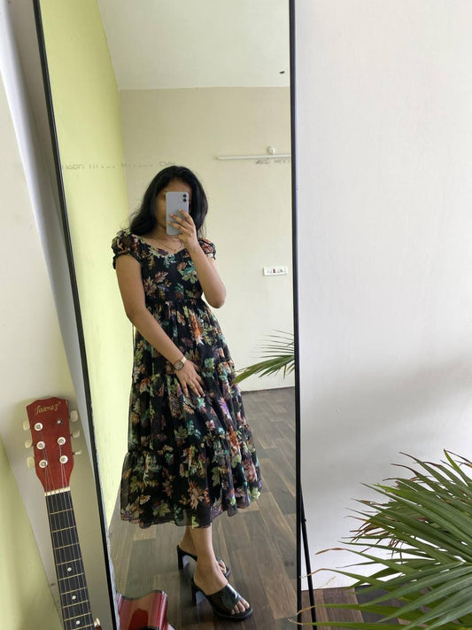 Woman in a black floral 'Floral Getaway' dress by a mirror. Faux georgette tiered dress with short sleeves.