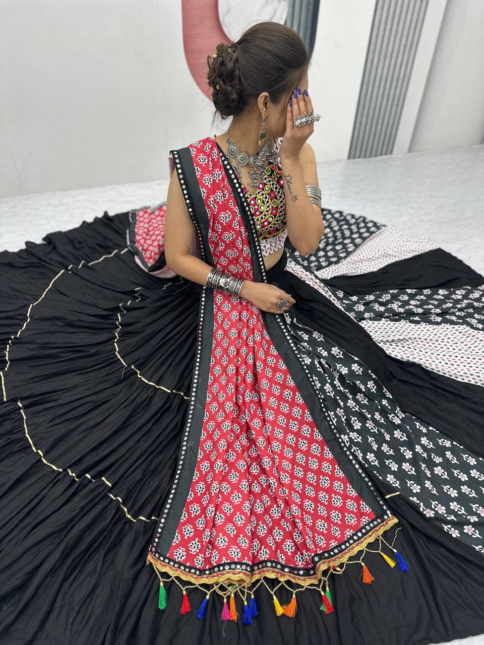 Model sitting in flared lehenga, showcasing multi-color panels and red dupatta with tassels.