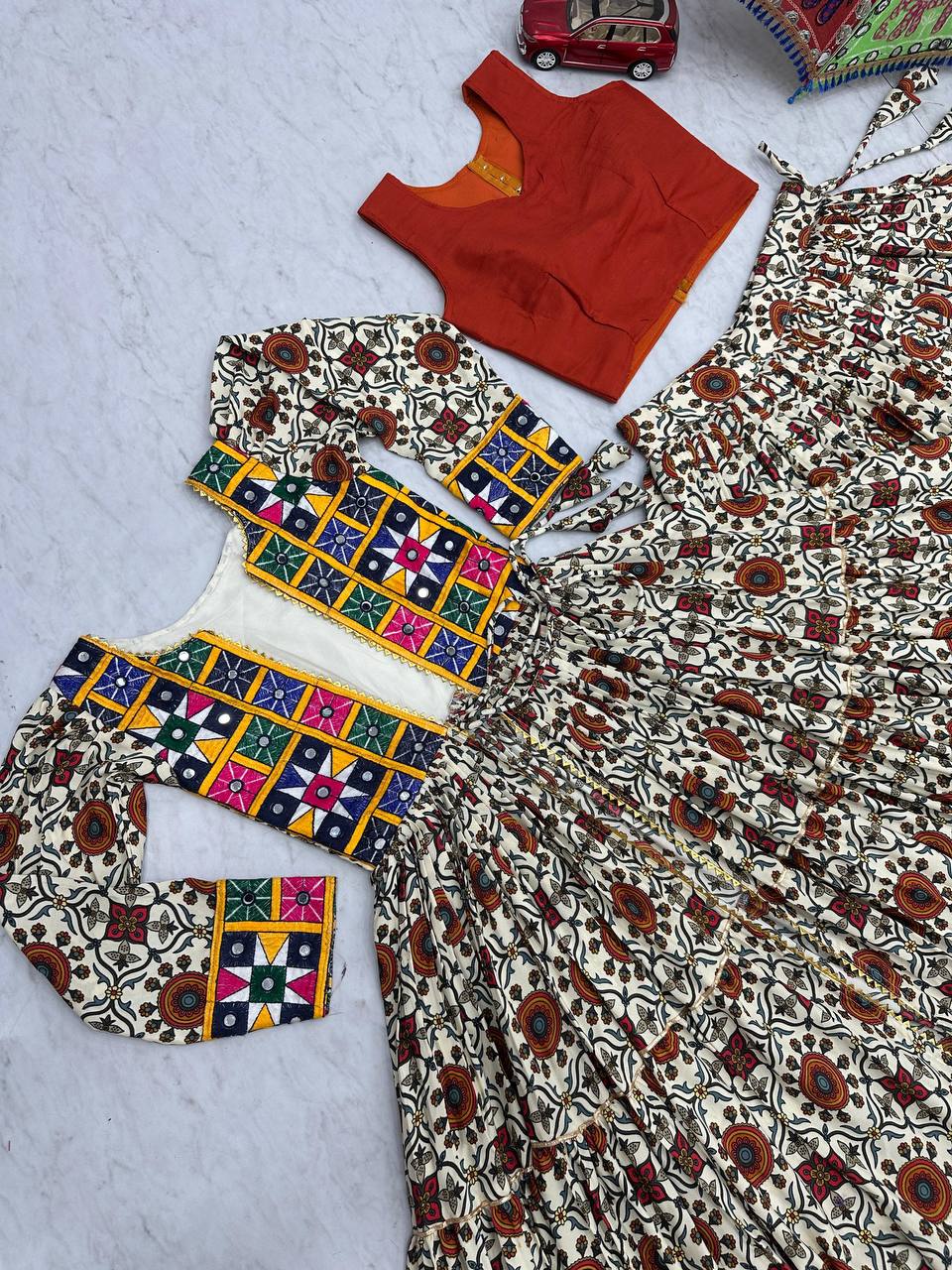 Flat lay of Kachchi Charm lehenga choli set showing orange blouse, mirror work shrug, and festive skirt.