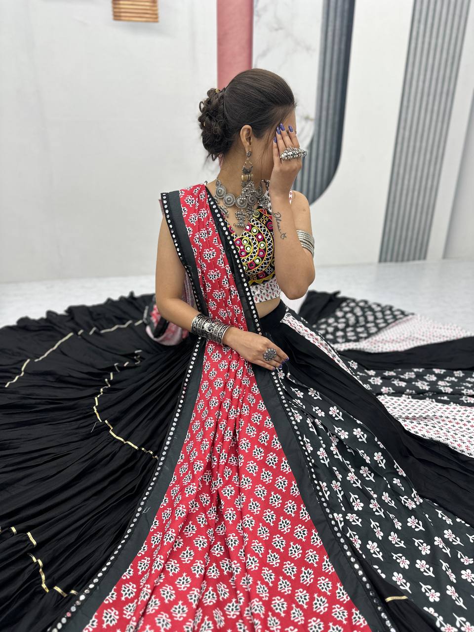 Side view of seated model wearing black, red, and white lehenga with festive dupatta and jewelry.
