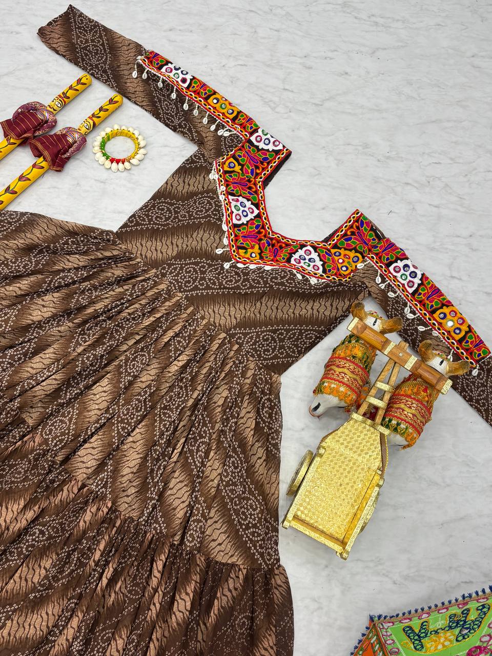 Brown Bandhni Gamathi work gown flat lay, with mirror work yoke and dandiya sticks for Navratri.