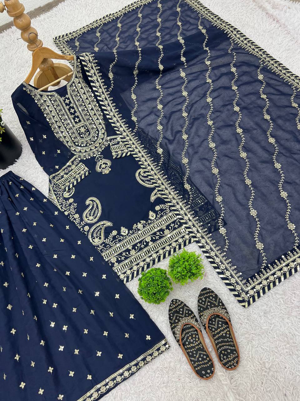 Flat lay of three-piece navy blue sharara set with gold embroidery, dupatta, and traditional juttis.
