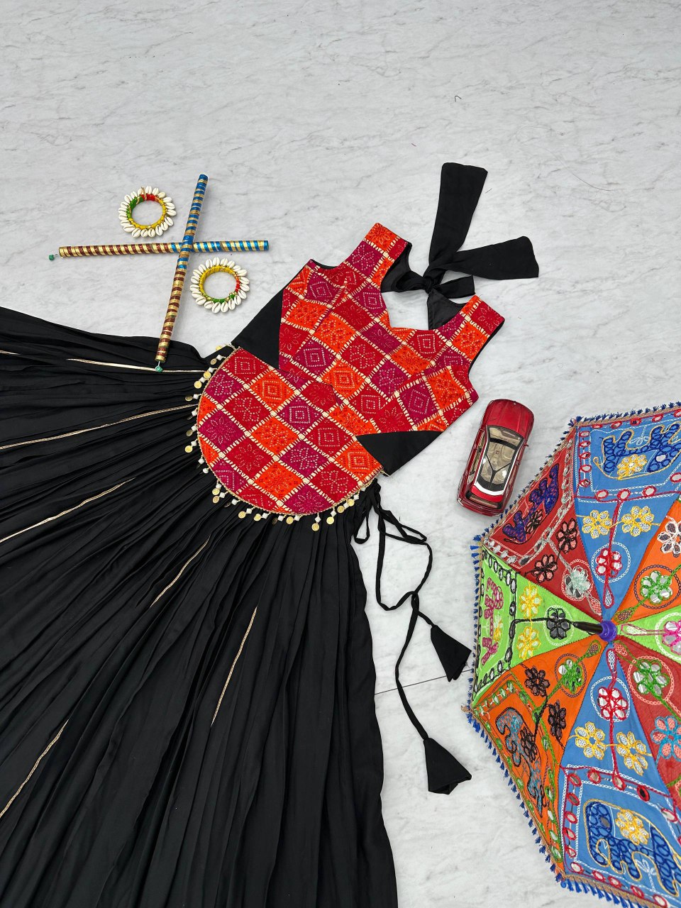 Flat lay of lehenga choli set with black skirt, red geometric choli, dandiya sticks and colorful props.