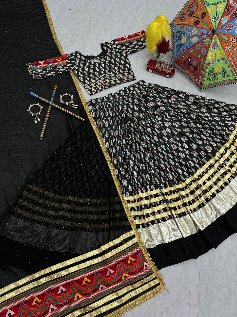 Flat lay: black lehenga choli set with gold border, ribbon work, dupatta, and festive accessories.
