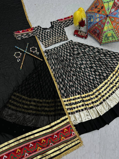 Flat lay: black lehenga choli set with gold border, ribbon work, dupatta, and festive accessories.