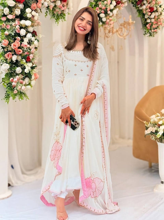 Model in off-white gown with gold embroidery and pink floral dupatta, smiling at event venue.