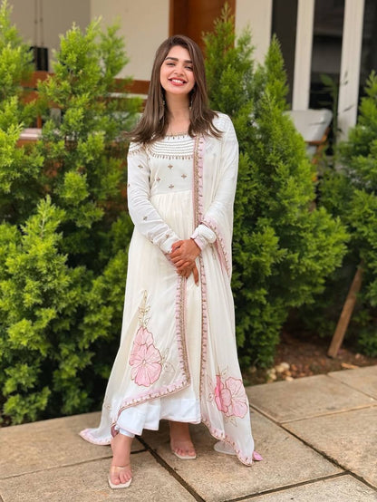 Woman in off-white embroidered gown with pastel pink floral dupatta, standing on patio.